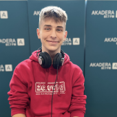 Piotr Tumiel, młody mężczyzna w czerwonej bluzie z logo „Zerówka”, siedzi uśmiechnięty w studiu Radia Akadera, na szyi ma słuchawki. W tle niebieskie ścianki z logo „AKADERA 87.7 FM”.