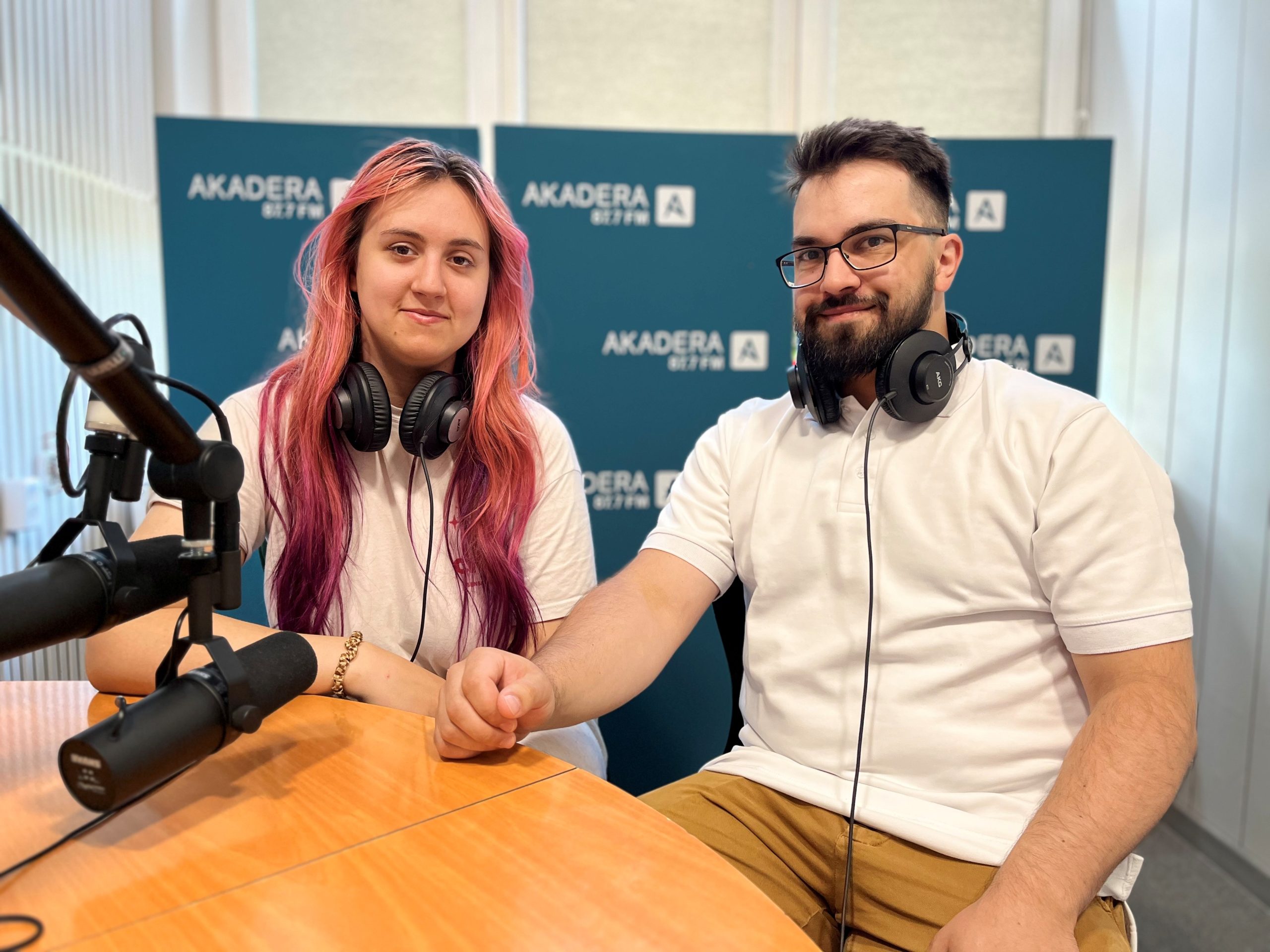 Ewa Tatarczyk i Szymon Podziewski siedzą przy stole radiowym w studiu Radia Akadera. Oboje mają na szyi słuchawki i uśmiechają się do obiektywu. W tle ścianka z logo Akadera 87.7 FM.