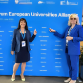 Dwie kobiety w niebieskich marynarkach stoją na tle niebieskiej ścianki z logo III Forum Uniwersytetów Europejskich we Wrocławiu i wskazują na nazwę swojego sojuszu ACROSS.