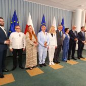 Na zdjęciu widoczna jest grupa osób nagrodzonych odznaczeniami państwowymi, stojących na tle flag Polski i Unii Europejskiej. Wydarzenie ma charakter oficjalny – widać odświętny ubiór uczestników oraz odznaczenia przypięte do klap marynarek i sukienek. Zdjęcie wykonano najpewniej po zakończeniu ceremonii wręczenia medali, w trakcie pamiątkowego zdjęcia z przedstawicielami władz. Gratulacje dla wszystkich uhonorowanych!