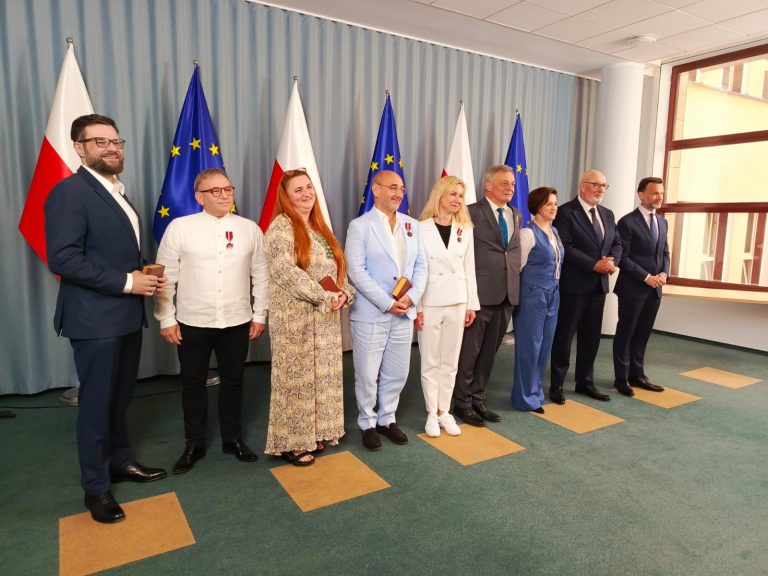 Na zdjęciu widoczna jest grupa osób nagrodzonych odznaczeniami państwowymi, stojących na tle flag Polski i Unii Europejskiej. Wydarzenie ma charakter oficjalny – widać odświętny ubiór uczestników oraz odznaczenia przypięte do klap marynarek i sukienek. Zdjęcie wykonano najpewniej po zakończeniu ceremonii wręczenia medali, w trakcie pamiątkowego zdjęcia z przedstawicielami władz. Gratulacje dla wszystkich uhonorowanych!
