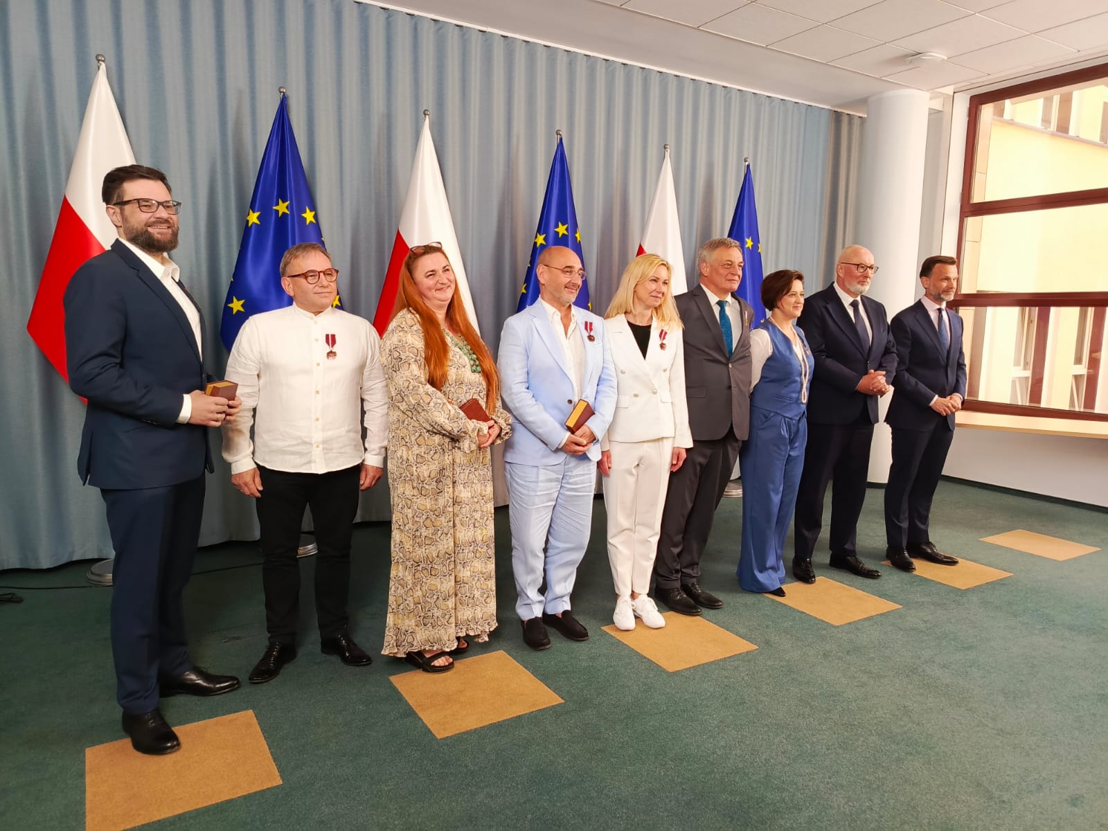 Na zdjęciu widoczna jest grupa osób nagrodzonych odznaczeniami państwowymi, stojących na tle flag Polski i Unii Europejskiej. Wydarzenie ma charakter oficjalny – widać odświętny ubiór uczestników oraz odznaczenia przypięte do klap marynarek i sukienek. Zdjęcie wykonano najpewniej po zakończeniu ceremonii wręczenia medali, w trakcie pamiątkowego zdjęcia z przedstawicielami władz. Gratulacje dla wszystkich uhonorowanych!