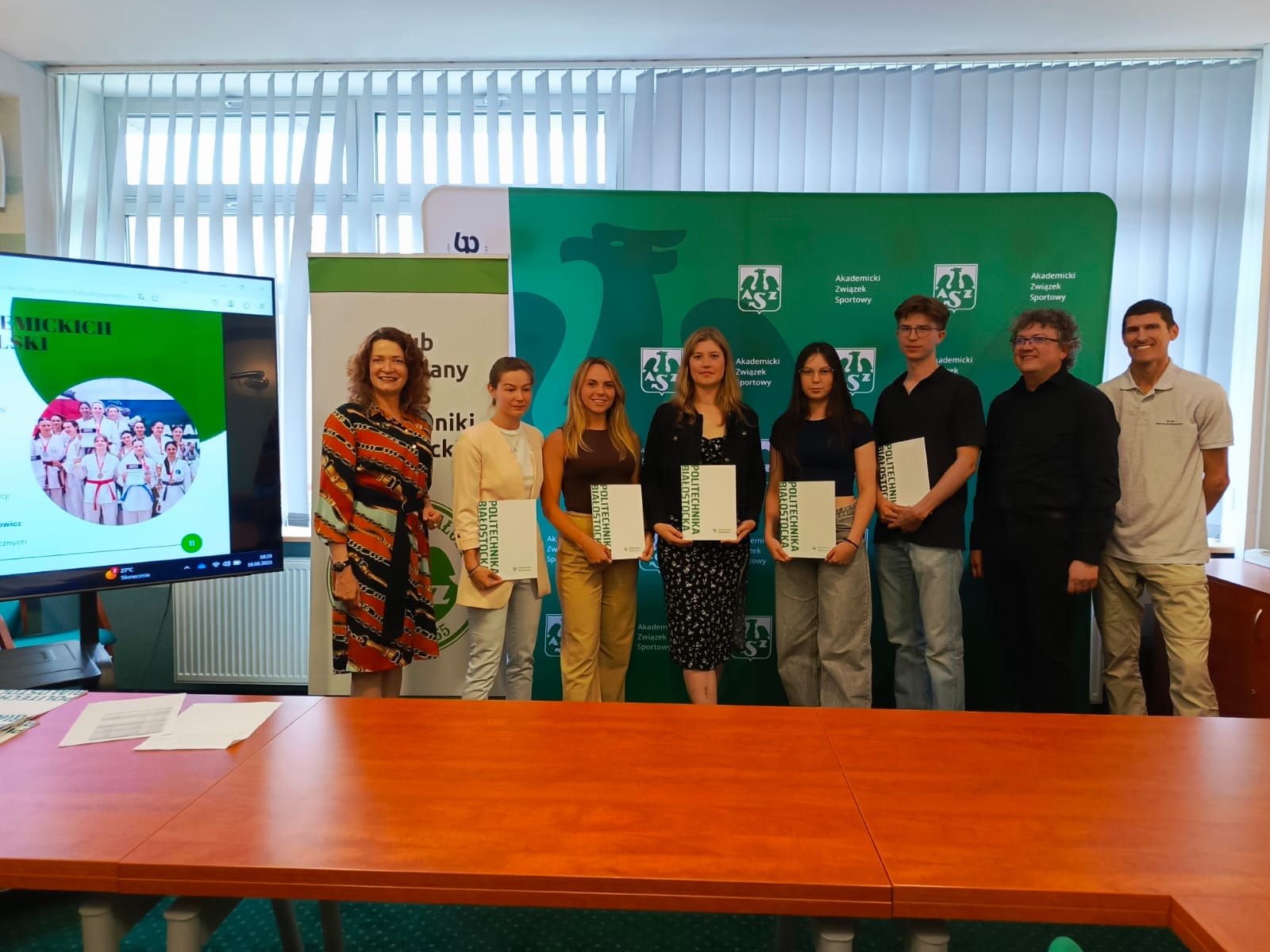 Na zdjęciu uwieczniono uroczystość zakończenia roku akademickiego w Akademickim Związku Sportowym Politechniki Białostockiej. Wyróżnieni studenci trzymają dyplomy, co świadczy o docenieniu ich sportowych osiągnięć. Obecni są również opiekunowie i przedstawiciele władz uczelni. Uroczysta atmosfera, uśmiechy i tło z logotypami AZS oraz PB podkreślają rangę wydarzenia i zaangażowanie społeczności akademickiej w rozwój sportu.