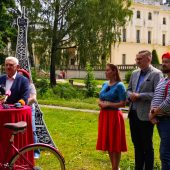 Zdjęcie przedstawia konferencję prasową lub wydarzenie promocyjne odbywające się na tle Pałacu Branickich w Białymstoku. Przy mikrofonach stoi mężczyzna w garniturze, obok którego znajdują się osoby w stylizacjach nawiązujących do francuskiej kultury – berety, paski, czerwone dodatki. W tle widoczna jest tekturowa makieta Wieży Eiffla oraz rower z koszem wypełnionym bagietkami, co sugeruje tematykę francuską wydarzenia – być może zapowiedź festiwalu kultury francuskiej, pikniku tematycznego lub innego wydarzenia promującego Francję w Białymstoku.