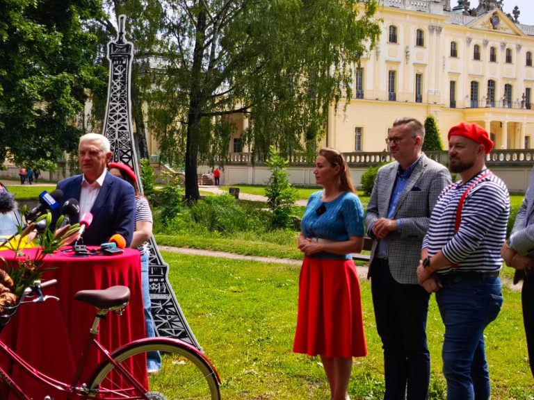 Zdjęcie przedstawia konferencję prasową lub wydarzenie promocyjne odbywające się na tle Pałacu Branickich w Białymstoku. Przy mikrofonach stoi mężczyzna w garniturze, obok którego znajdują się osoby w stylizacjach nawiązujących do francuskiej kultury – berety, paski, czerwone dodatki. W tle widoczna jest tekturowa makieta Wieży Eiffla oraz rower z koszem wypełnionym bagietkami, co sugeruje tematykę francuską wydarzenia – być może zapowiedź festiwalu kultury francuskiej, pikniku tematycznego lub innego wydarzenia promującego Francję w Białymstoku.