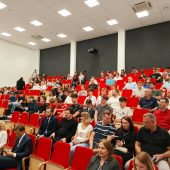 Na zdjęciu widzimy pełną widownię zgromadzoną w nowoczesnej auli z czerwonymi fotelami. Uczestnicy – głównie młodzież, ale też nauczyciele, rodzice i przedstawiciele uczelni – biorą udział w wydarzeniu, najprawdopodobniej związanym z konkursem „Invent Your Game 2025”. Panuje atmosfera skupienia i oczekiwania, co sugeruje, że wydarzenie dopiero się rozpoczyna lub trwa jego oficjalna część.