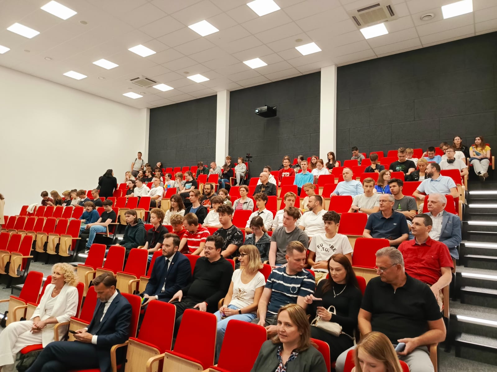 Na zdjęciu widzimy pełną widownię zgromadzoną w nowoczesnej auli z czerwonymi fotelami. Uczestnicy – głównie młodzież, ale też nauczyciele, rodzice i przedstawiciele uczelni – biorą udział w wydarzeniu, najprawdopodobniej związanym z konkursem „Invent Your Game 2025”. Panuje atmosfera skupienia i oczekiwania, co sugeruje, że wydarzenie dopiero się rozpoczyna lub trwa jego oficjalna część.