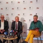 Na zdjęciu widać konferencję prasową zorganizowaną przez Teatr Dramatyczny im. Aleksandra Węgierki w Białymstoku. Na tle ścianki z logo teatru i Województwa Podlaskiego siedzi trzech mężczyzn: po lewej – mężczyzna w garniturze z białą koszulą i okularami, w środku – mężczyzna w garniturze i krawacie, udzielający wypowiedzi do zgromadzonych mediów, a po prawej – mężczyzna w zielonej bluzie i żółtych spodniach, trzymający dokumenty. Przed nimi znajduje się stolik z mikrofonami i rejestratorami należącymi do lokalnych mediów, m.in. TVP3 Białystok, Polskiego Radia Białystok i Radia Białystok. Atmosfera sugeruje oficjalne podsumowanie wydarzenia lub zapowiedź nowych inicjatyw teatralnych.