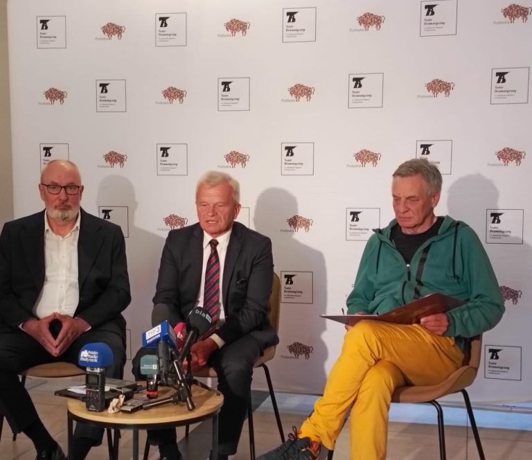 Na zdjęciu widać konferencję prasową zorganizowaną przez Teatr Dramatyczny im. Aleksandra Węgierki w Białymstoku. Na tle ścianki z logo teatru i Województwa Podlaskiego siedzi trzech mężczyzn: po lewej – mężczyzna w garniturze z białą koszulą i okularami, w środku – mężczyzna w garniturze i krawacie, udzielający wypowiedzi do zgromadzonych mediów, a po prawej – mężczyzna w zielonej bluzie i żółtych spodniach, trzymający dokumenty. Przed nimi znajduje się stolik z mikrofonami i rejestratorami należącymi do lokalnych mediów, m.in. TVP3 Białystok, Polskiego Radia Białystok i Radia Białystok. Atmosfera sugeruje oficjalne podsumowanie wydarzenia lub zapowiedź nowych inicjatyw teatralnych.