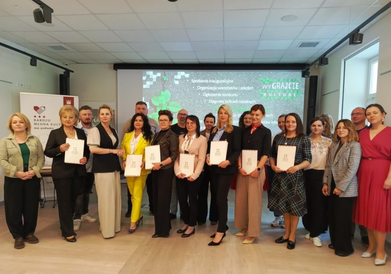 kobiety i mezczyzni stoja prezentujac dokumenty w dloniach, pozuja do zdjecia na tle napisow wyswietlanych w tle na scianie
