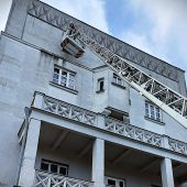 Strażak na wysięgniku drabiny ewakuacyjnej podczas akcji ratowniczej w hostelu przy ulicy Abramowicza w Białymstoku – budynek o jasnej elewacji, niebo z lekkim zachmurzeniem.