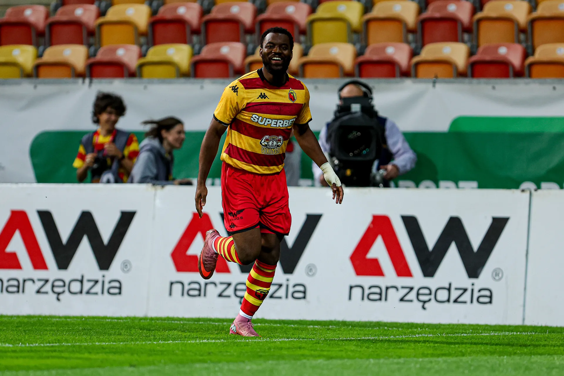 Afimico Pululu, piłkarz Jagiellonii Białystok w żółto-czerwonym stroju, uśmiechnięty biegnie po boisku podczas meczu, w tle trybuny i fotografowie.