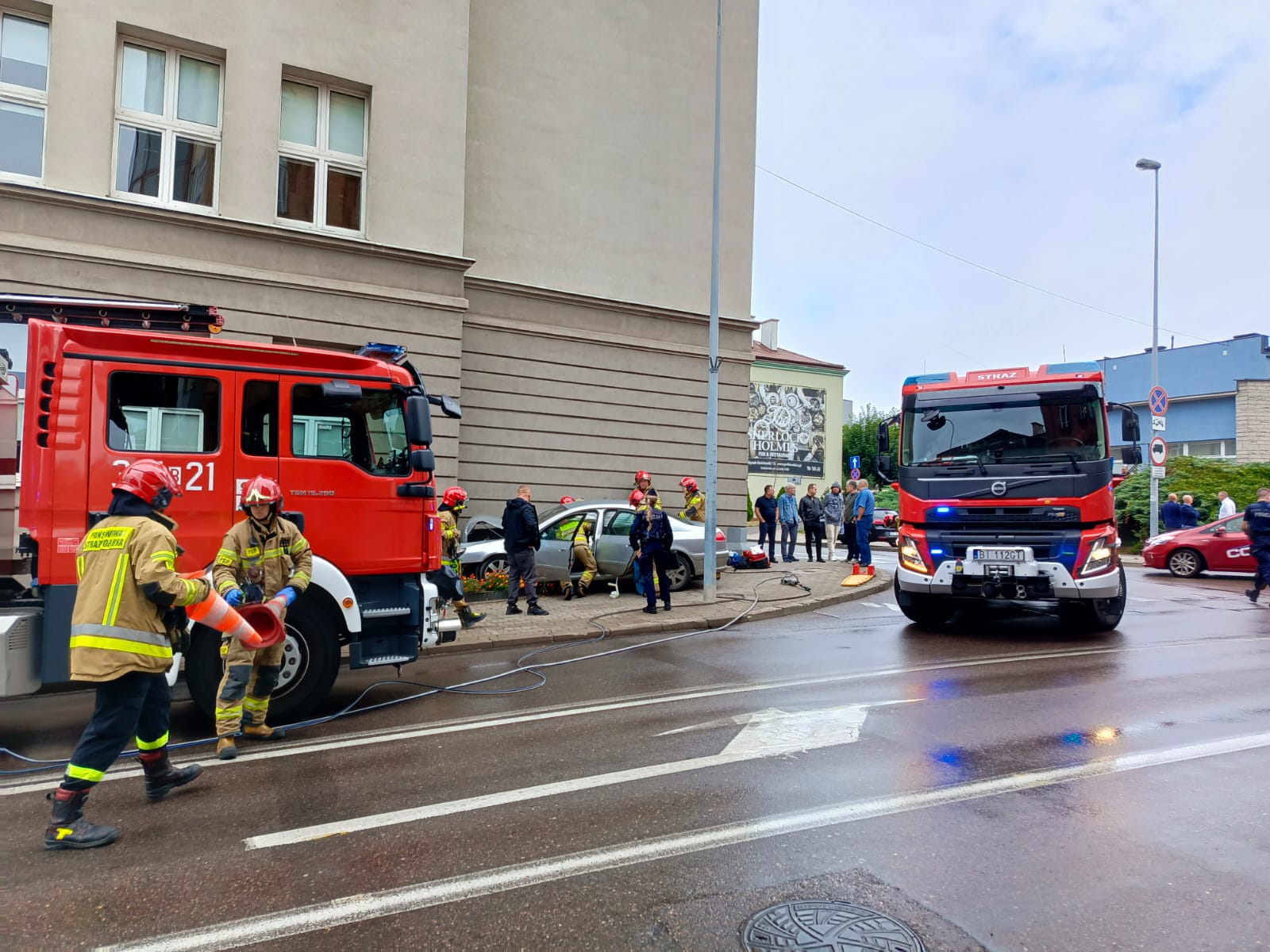 Strażacy i wozy straży pożarnej stoją przy rozbitym samochodzie, który uderzył w budynek III LO w Białymstoku.