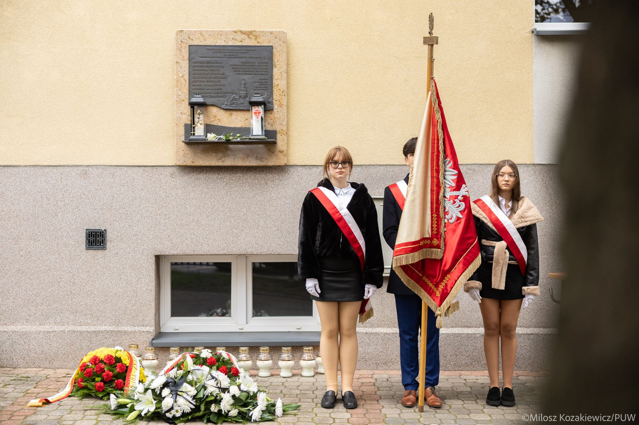 Troje uczniów pocztu sztandarowego I LO w Białymstoku stoi pod tablicą pamiątkową ofiar katastrofy autokaru pod Jeżewem. Obok widoczne są kwiaty i zapalone znicze.