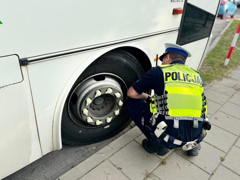 Policjant w odblaskowej kamizelce sprawdza stan techniczny autobusu szkolnego, skupiając się na kole pojazdu.