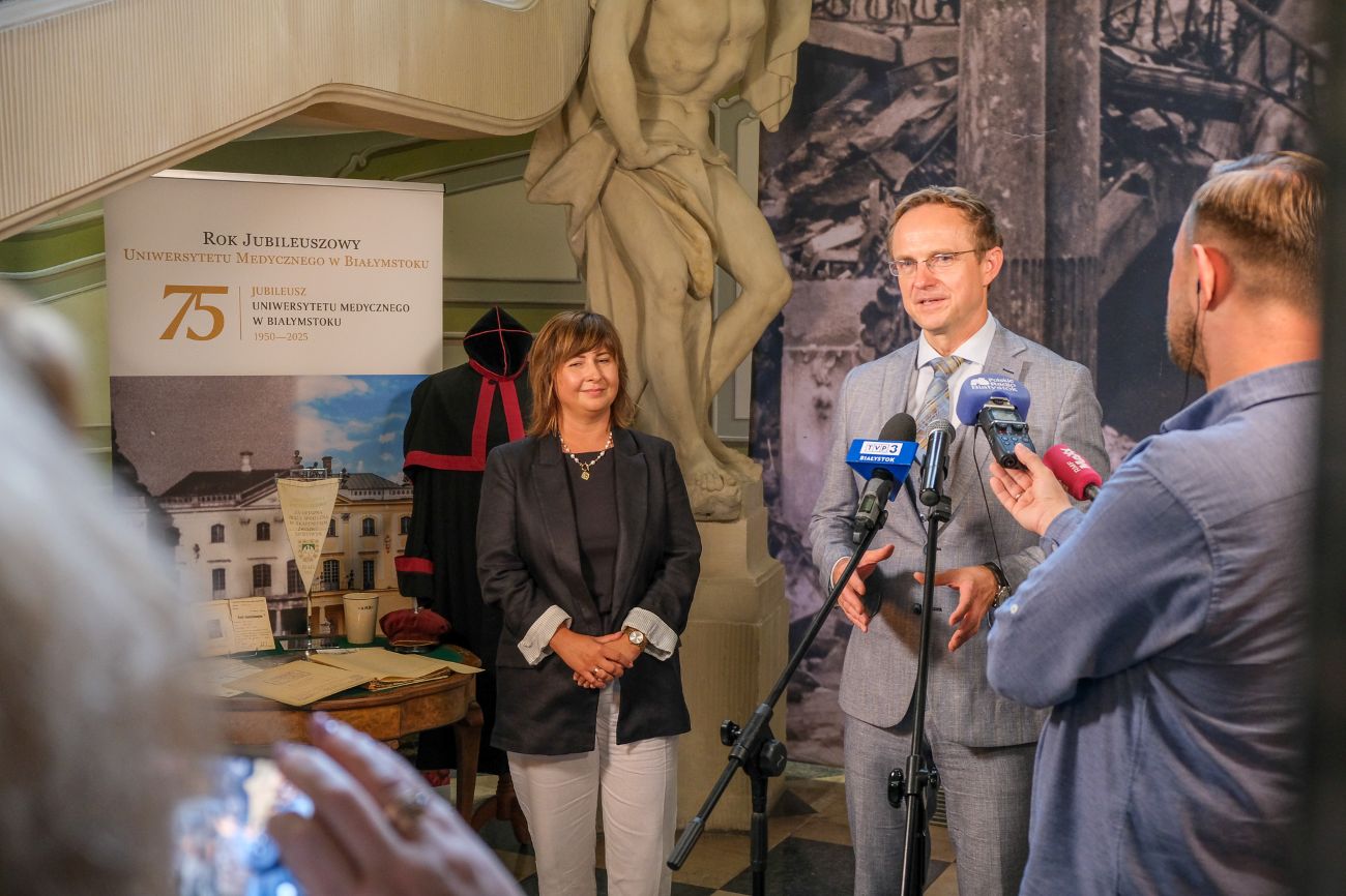 Rektor Uniwersytetu Medycznego w Białymstoku prof. Marcin Moniuszko przemawia na konferencji prasowej; obok stoi dr Magdalena Muskała, dyrektor Muzeum UMB i Pałacu Branickich. W tle widoczny baner jubileuszu 75-lecia uczelni i elementy ekspozycji muzealnej.