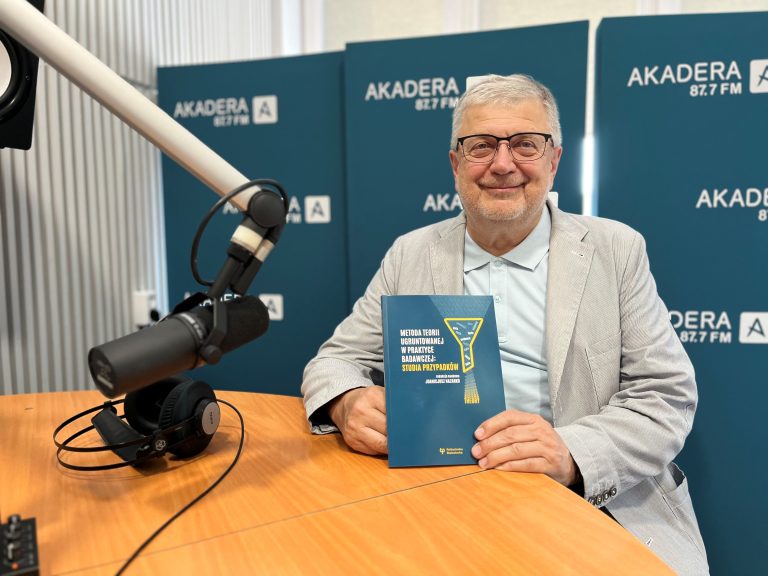 Profesor Joanicjusz Nazarko siedzi w studiu Radia Akadera, trzymając w dłoniach książkę „Metoda teorii ugruntowanej w praktyce badawczej: studia przypadków” na tle ścianki z logo Radia Akadera.