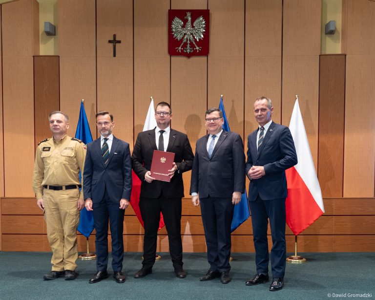 Wojewoda podlaski Jacek Brzozowski przekazuje zastępcy prezydenta Białegostoku Tomaszowi Klimowi dokument potwierdzający przyznanie dotacji na ochronę ludności. Na zdjęciu pięciu mężczyzn w oficjalnych strojach podczas uroczystości w urzędzie, na tle flag Polski i UE.