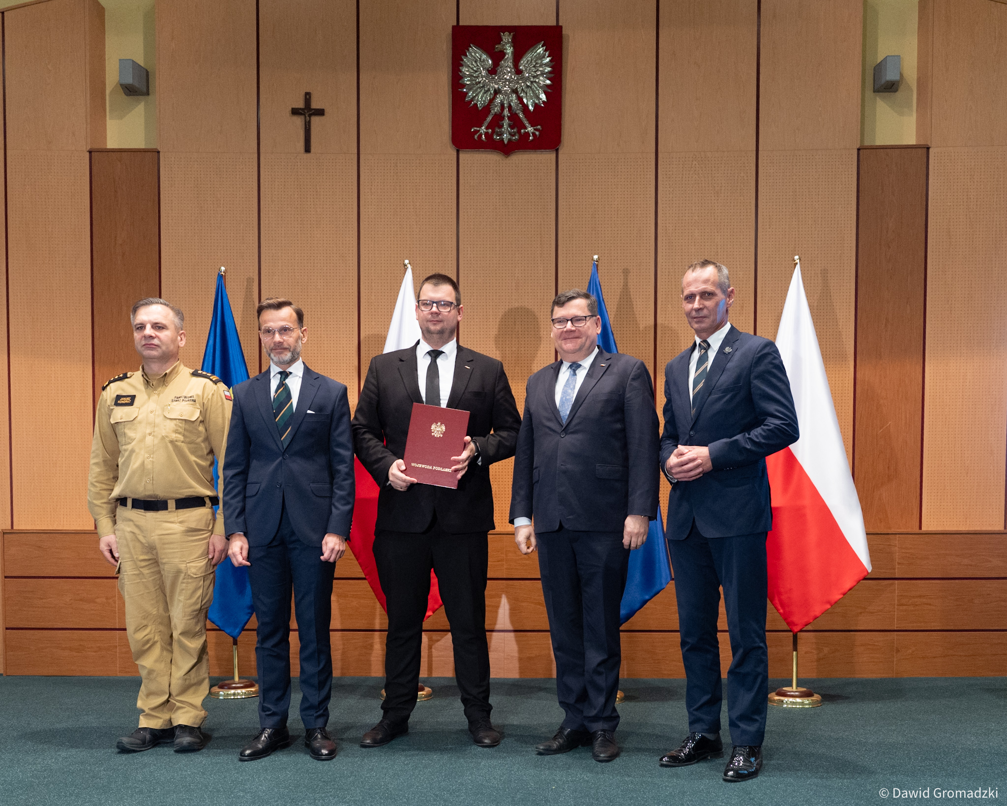 Wojewoda podlaski Jacek Brzozowski przekazuje zastępcy prezydenta Białegostoku Tomaszowi Klimowi dokument potwierdzający przyznanie dotacji na ochronę ludności. Na zdjęciu pięciu mężczyzn w oficjalnych strojach podczas uroczystości w urzędzie, na tle flag Polski i UE.