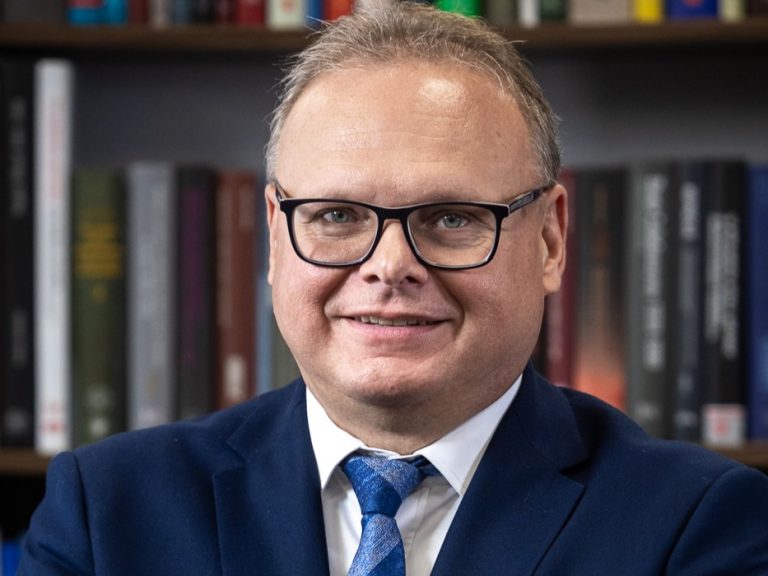 Portret mężczyzny w garniturze i okularach, uśmiechniętego, na tle półki z książkami. To dr hab. Krzysztof Sychowicz, nowy dyrektor Oddziału IPN w Białymstoku.