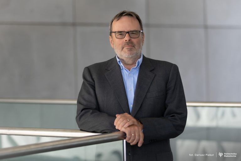 Prof. Krzysztof Kurzydłowski, Dyrektor Instytutu Inżynierii Mechanicznej Politechniki Białostockiej, stoi przy balustradzie w nowoczesnym wnętrzu uczelni. Ubrany w ciemny garnitur i niebieską koszulę, spogląda w stronę obiektywu.