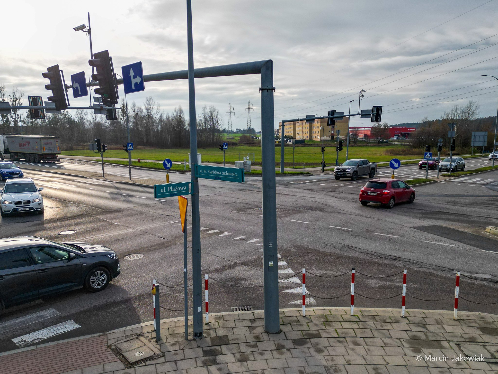 Skrzyżowanie ulic Suchowolca i Plażowej w Białymstoku, widok na sygnalizację świetlną, znaki drogowe i jadące samochody. Na zdjęciu widać nowe oznakowanie oraz układ pasów wprowadzany w ramach zmian organizacji ruchu.