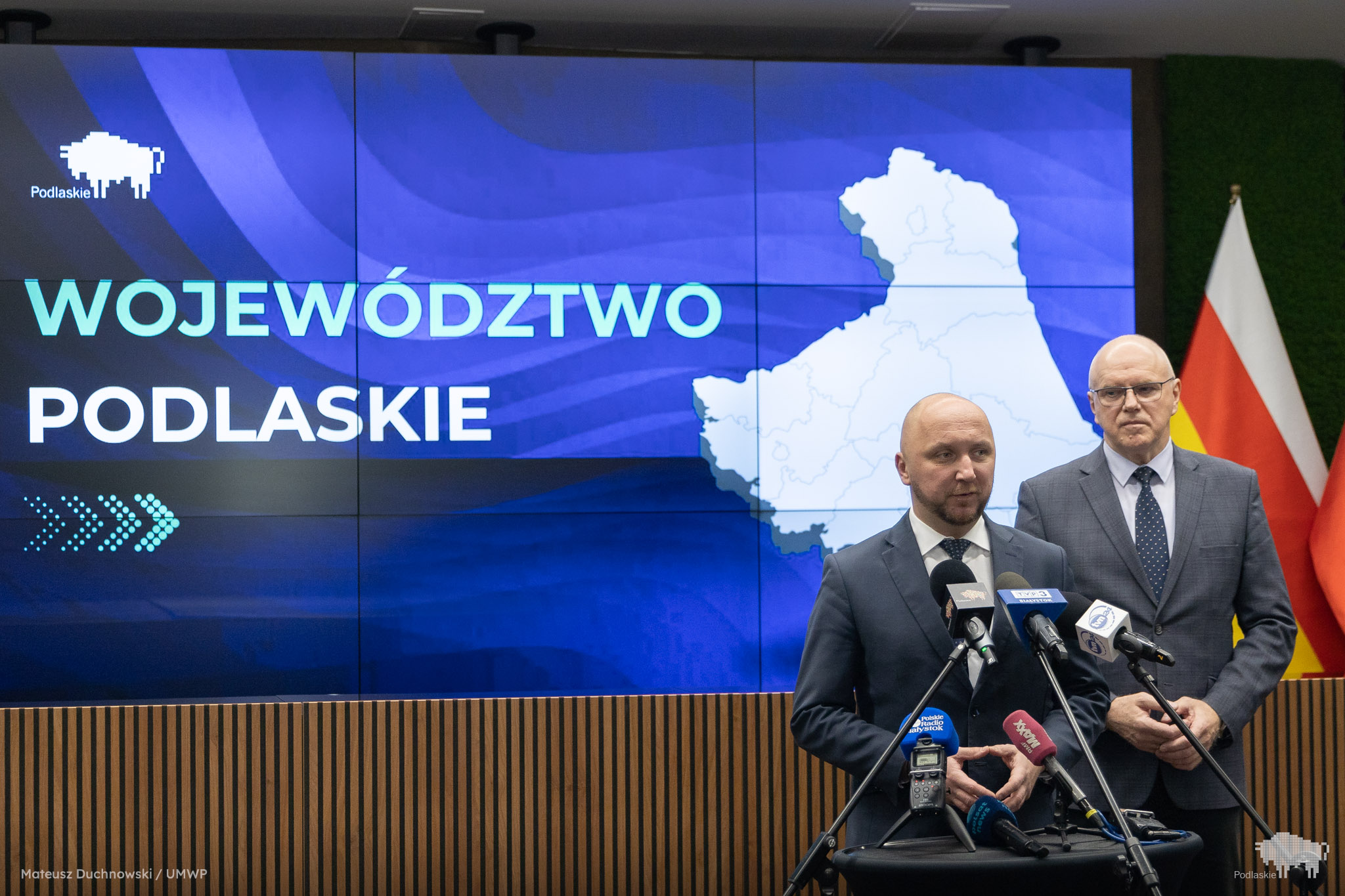 Marszałek Województwa Podlaskiego Łukasz Prokorym stoi przed mikrofonami podczas konferencji prasowej. Za nim widać członka zarządu województwa oraz duży ekran z mapą regionu i napisem „Województwo Podlaskie”.
