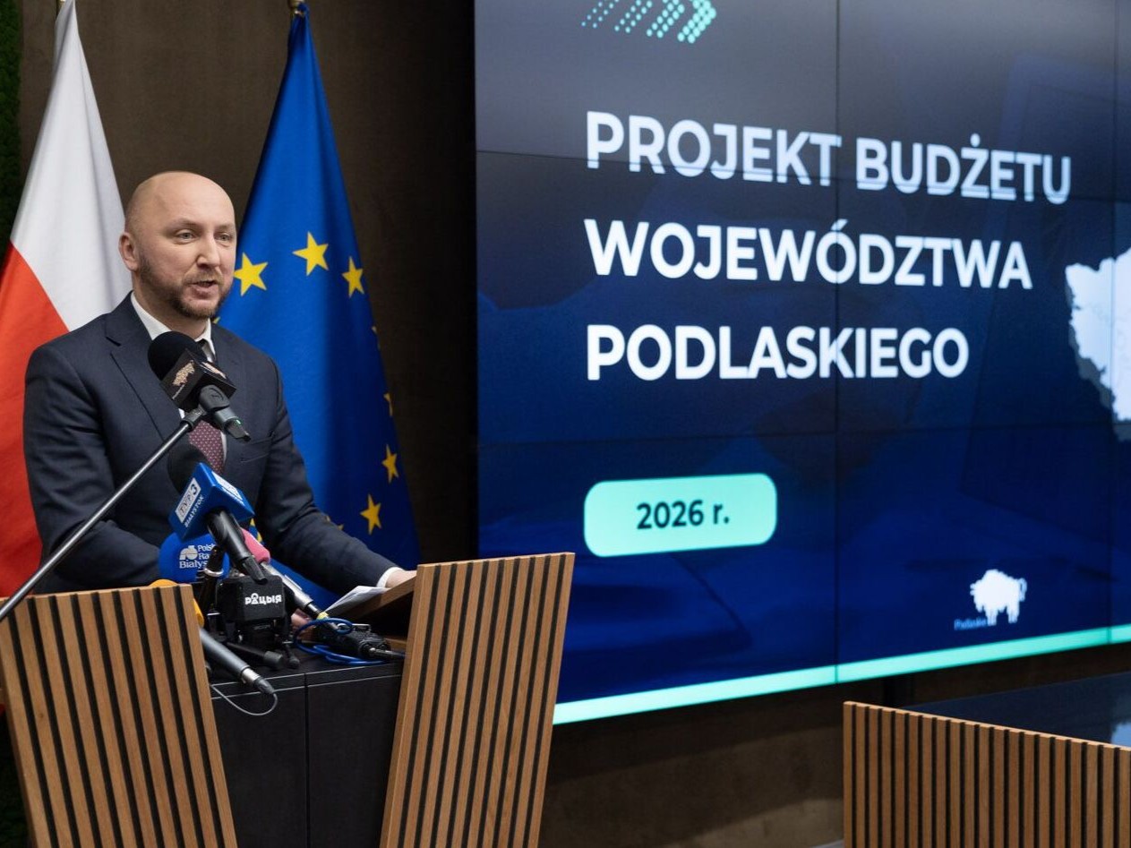 Marszałek Łukasz Prokorym stoi przy mównicy podczas konferencji prasowej, na tle ekranu z napisem „Projekt budżetu Województwa Podlaskiego 2026 r.” i mapą regionu; obok widoczne są flagi Polski, województwa podlaskiego i Unii Europejskiej.