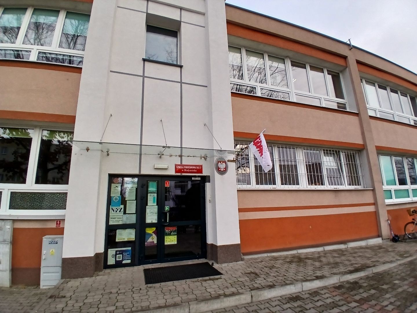 Wejście do Szkoły Podstawowej w Białymstoku z wywieszoną flagą NSZZ „Solidarność”. Na budynku widoczne są tablice informacyjne oraz godło państwowe.