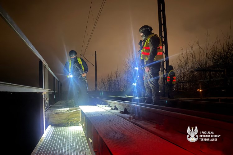 Żołnierze 1. Podlaskiej Brygady Obrony Terytorialnej patrolują nocą tory kolejowe z latarkami, stojąc na mostowej konstrukcji; w tle widoczne słupy trakcyjne i ciemne niebo.