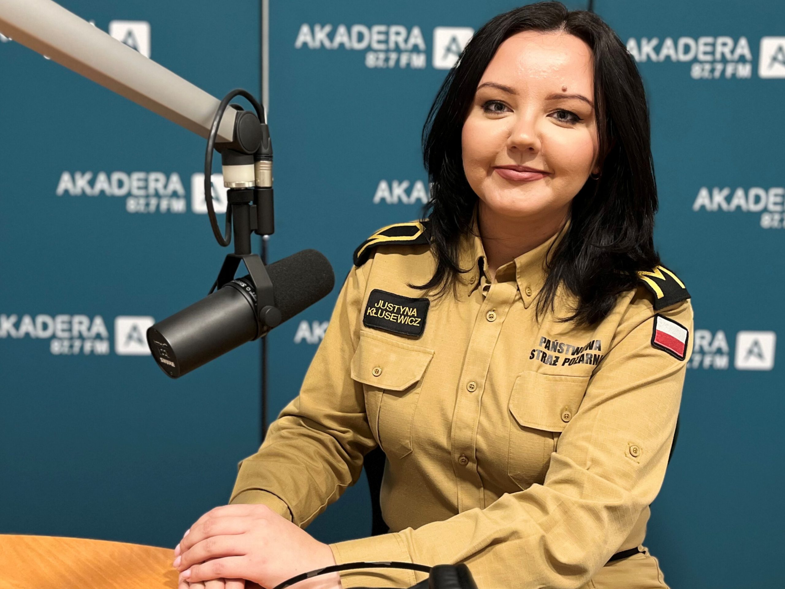 Ogniomistrz Justyna Kłusewicz, rzeczniczka prasowa Komendanta Wojewódzkiego PSP w Białymstoku, siedzi przy stole w studiu Radia Akadera. Przed nią stoi mikrofon i leżą słuchawki. W tle widoczne są niebieskie ścianki z logo „Akadera 87,7 FM”.