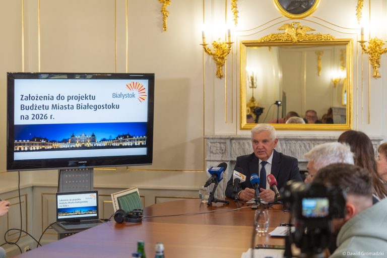 Prezydent Białegostoku Tadeusz Truskolaski siedzi przy stole konferencyjnym w sali Pałacu Branickich i przedstawia założenia budżetu miasta na 2026 rok. Obok ekranu z napisem „Założenia do projektu Budżetu Miasta Białegostoku na 2026 r.” stoją mikrofony lokalnych mediów.