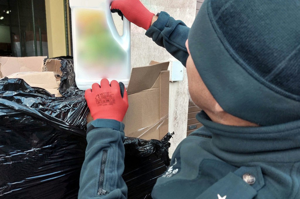 Funkcjonariusz KAS w czerwonych rękawicach ogląda pięciolitrową butelkę detergentu wyjętą z kartonu podczas kontroli transportu podejrzanego o przewóz podrobionych płynów do prania.