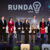 Siedmioro uczestników gali konkursu Runda T 2025 stoi na scenie przed dużym ekranem z logo wydarzenia. Wicemarszałek Marek Malinowski przemawia, trzymając mikrofon. Obok niego stoją przedstawiciele Fundacji Technotalenty oraz finaliści konkursu. Tło zdobi grafika żarówki symbolizująca innowacje.