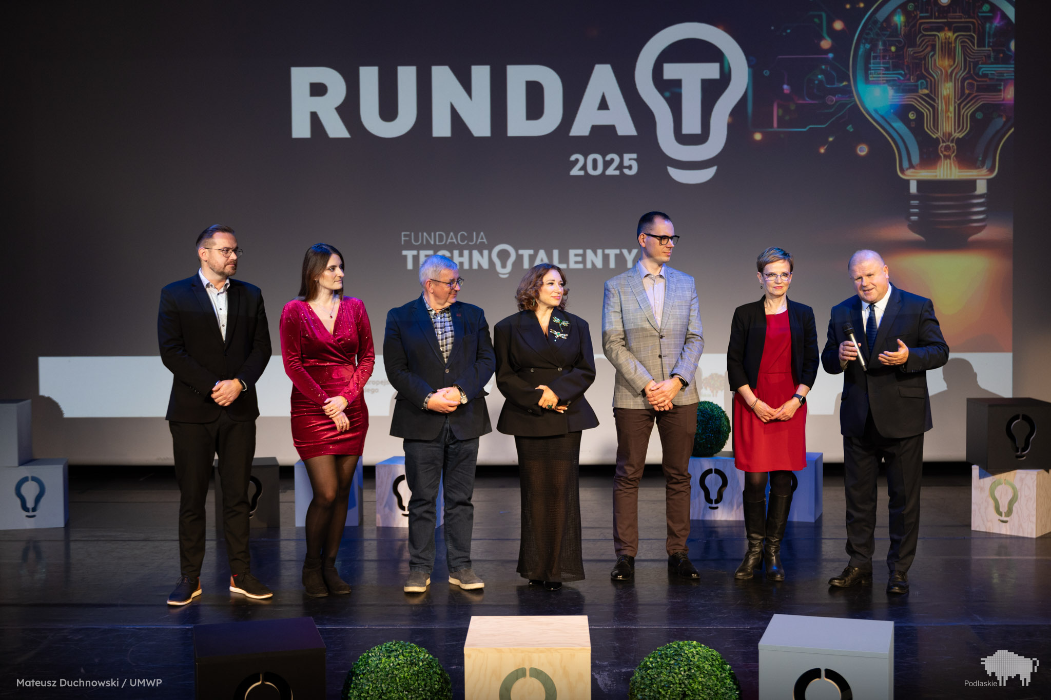 Siedmioro uczestników gali konkursu Runda T 2025 stoi na scenie przed dużym ekranem z logo wydarzenia. Wicemarszałek Marek Malinowski przemawia, trzymając mikrofon. Obok niego stoją przedstawiciele Fundacji Technotalenty oraz finaliści konkursu. Tło zdobi grafika żarówki symbolizująca innowacje.