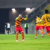 Piłkarze Jagiellonii Białystok w żółto-czerwonych strojach podczas meczu z GKS Katowice w 1/8 finału Pucharu Polski. Zawodnik na pierwszym planie unosi zaciśnięte pięści w geście mobilizacji, a obok niego widoczny jest afimico Pululu z numerem 10 na koszulce. W tle fragment stadionu i pozostali piłkarze.