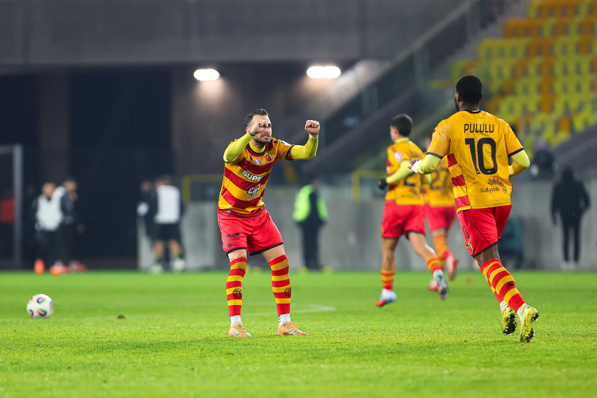 Piłkarze Jagiellonii Białystok w żółto-czerwonych strojach podczas meczu z GKS Katowice w 1/8 finału Pucharu Polski. Zawodnik na pierwszym planie unosi zaciśnięte pięści w geście mobilizacji, a obok niego widoczny jest afimico Pululu z numerem 10 na koszulce. W tle fragment stadionu i pozostali piłkarze.