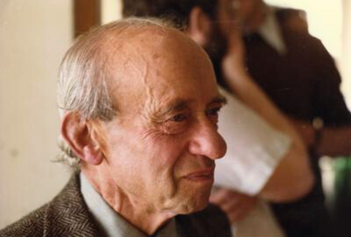 Kolorowa fotografia Alfreda Tarskiego w podeszłym wieku. Uśmiechnięty mężczyzna o siwych włosach, ubrany w marynarkę i koszulę z krawatem. W tle półki z książkami w jego gabinecie w Berkeley.