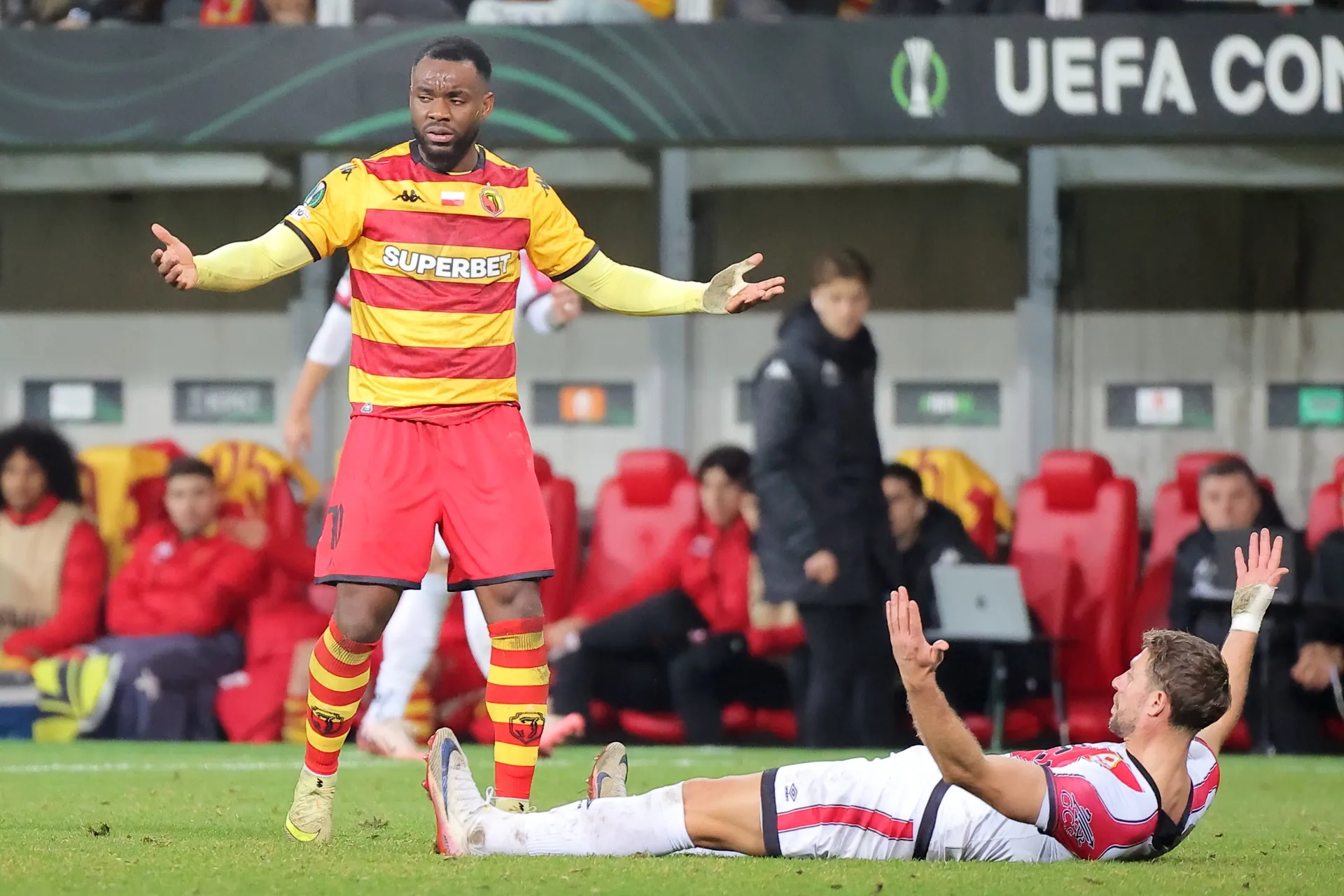 Piłkarz Jagiellonii Białystok gestykuluje z rozłożonymi rękami obok leżącego na murawie zawodnika Rayo Vallecano podczas meczu Ligi Konferencji na stadionie w Białymstoku.