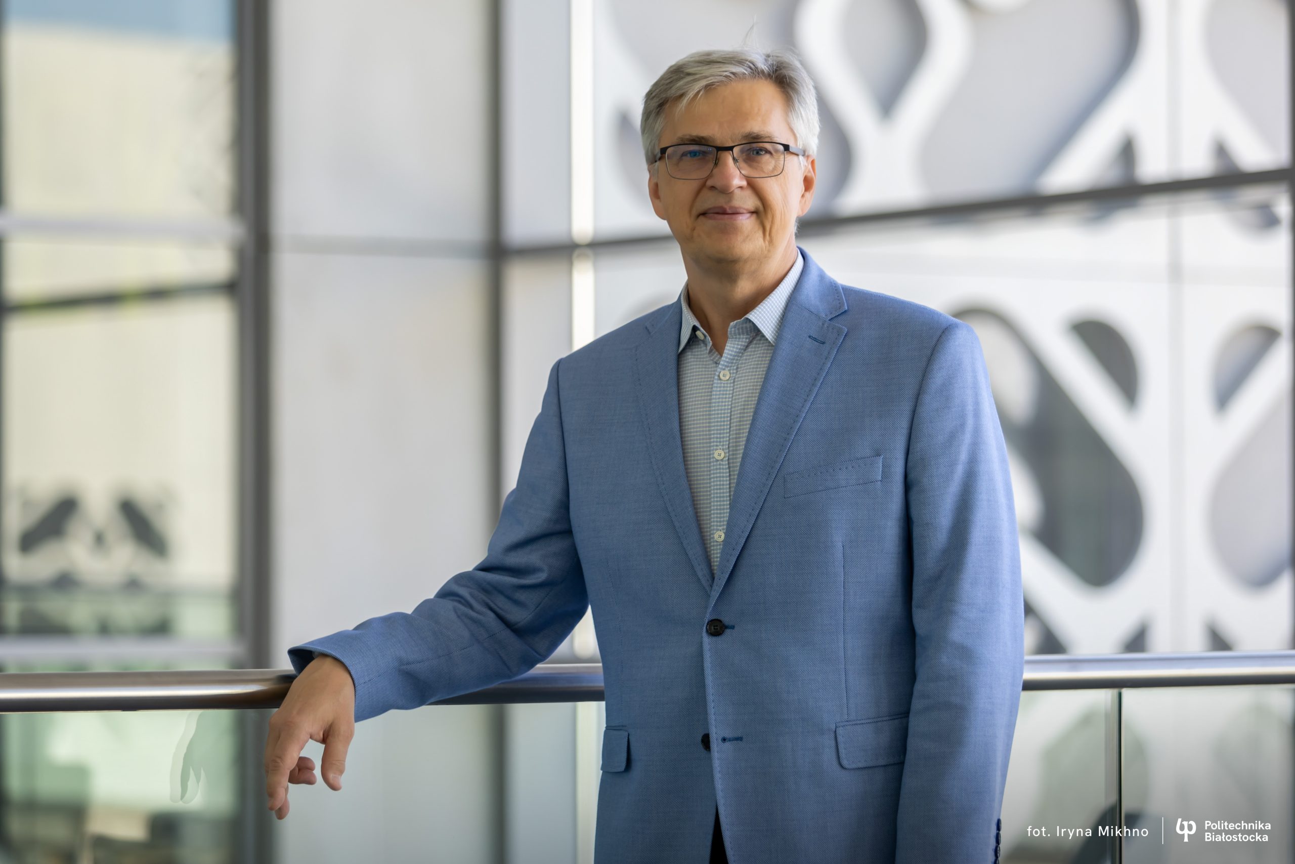 Dr hab. inż. Bogusław Butryło, prof. PB, dziekan Wydziału Elektrycznego Politechniki Białostockiej, stoi w nowoczesnym wnętrzu uczelni, opierając się o balustradę.